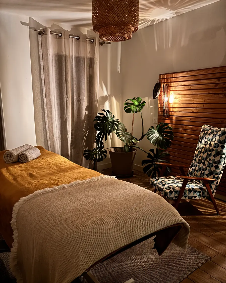 La table de massage préparée, son plaid de lin et ses serviettes roulées, devant le monstera et le mur de bois, à la lumière du soir